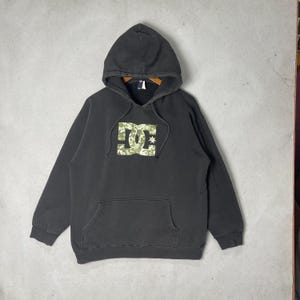 May include: A charcoal gray hooded sweatshirt featuring a large DC logo on the chest. The logo is filled with a light green and white camouflage pattern. The hoodie includes a front kangaroo pocket, long sleeves, and matching drawstring cords.