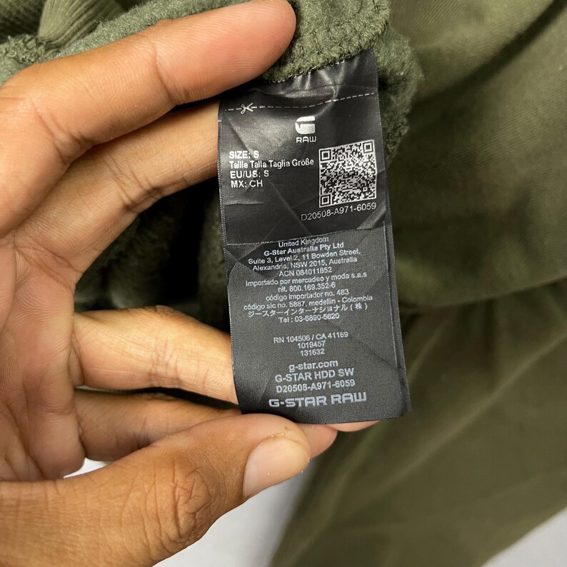 Puede incluir: Primer plano de una etiqueta negra de ropa G-Star Raw. La etiqueta incluye la marca, la talla y la informaci&oacute;n del producto. La etiqueta est&aacute; sujeta por una mano y el fondo es una tela verde.