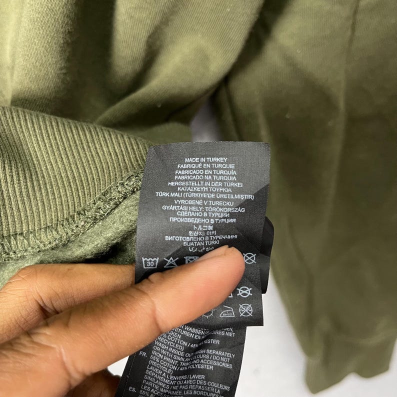 Puede incluir: Primer plano de un pu&ntilde;o de sudadera verde. Se muestra una etiqueta negra con texto blanco, que muestra las instrucciones de lavado y la frase "MADE IN TURKEY" en varios idiomas. La tela es de color verde oliva liso.