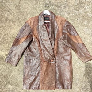 May include: Brown leather jacket with a textured design. The jacket has a wide collar, two buttons, and long sleeves. The jacket is hanging on a white hanger.