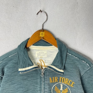 Peut inclure: Veste zipp&eacute;e bleu sarcelle avec bordure blanche et int&eacute;rieur cr&egrave;me. La veste pr&eacute;sente un &eacute;cusson "Air Force" et un embl&egrave;me dor&eacute;. La marque "AVIREX" est visible sur une &eacute;tiquette. La veste est suspendue &agrave; un cintre en bois.