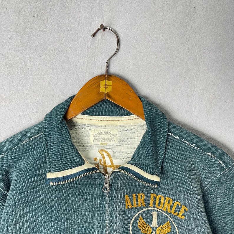 Peut inclure: Veste zipp&eacute;e bleu sarcelle avec bordure blanche et int&eacute;rieur cr&egrave;me. La veste pr&eacute;sente un &eacute;cusson "Air Force" et un embl&egrave;me dor&eacute;. La marque "AVIREX" est visible sur une &eacute;tiquette. La veste est suspendue &agrave; un cintre en bois.