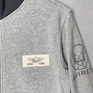 以下が含まれることがあります： ダークリブカラーが付いたヘザーグレーのジップアップジャケット。胸元の長方形のパッチには、851BSおよび490BGというテキストと共に翼の付いた爆弾のグラフィックが描かれています。袖には翼、星、AVIREXというテキストが入った円形のロゴがあります。
