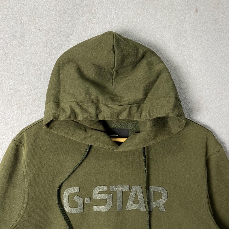 Puede incluir: Sudadera con capucha verde oliva con capucha con cord&oacute;n y el logotipo de G-Star en el pecho. La sudadera con capucha est&aacute; hecha de un material suave y tiene un ajuste relajado. El fondo es gris neutro.