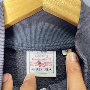 Puede incluir: Primer plano de una sudadera gris con cremallera con una etiqueta que dice "Certificate of Authenticity" y "AVIREX U.S.A.". La etiqueta tambi&eacute;n muestra la bandera estadounidense y la talla "M". La sudadera est&aacute; colgada en una percha de madera.