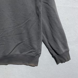 Puede incluir: Primer plano de una sudadera gris oscuro. La sudadera tiene un dobladillo y pu&ntilde;os acanalados. La tela parece ser un material suave y desgastado. La manga es visible, con el pu&ntilde;o en la parte inferior.