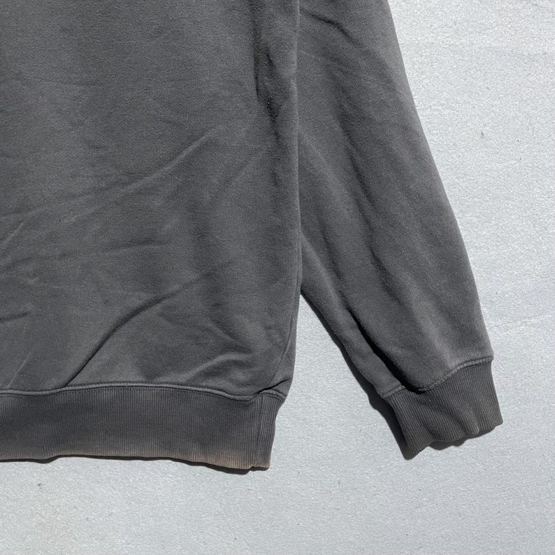 Puede incluir: Primer plano de una sudadera gris oscuro. La sudadera tiene un dobladillo y pu&ntilde;os acanalados. La tela parece ser un material suave y desgastado. La manga es visible, con el pu&ntilde;o en la parte inferior.