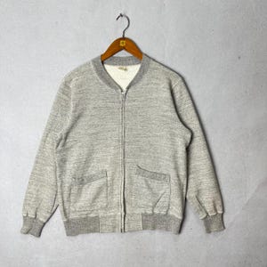 May include: A gray zip-up jacket with two front pockets and a ribbed collar and cuffs. The jacket is hanging on a wooden hanger against a gray background. The jacket is made of a soft, textured material.