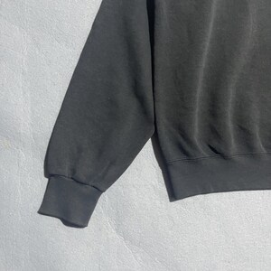 Op de afbeelding: Een close-up van een zwarte sweatshirtmouw. De mouw heeft een geribbelde manchet en is gemaakt van een zacht, licht vervaagd materiaal. Het sweatshirt lijkt versleten, met subtiele variaties in de textuur van de stof.