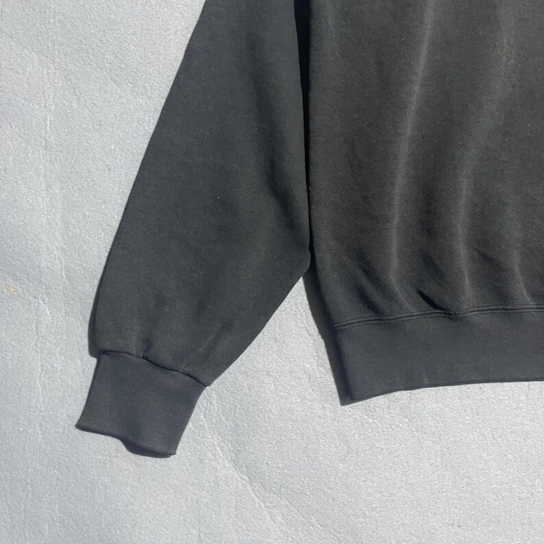 Op de afbeelding: Een close-up van een zwarte sweatshirtmouw. De mouw heeft een geribbelde manchet en is gemaakt van een zacht, licht vervaagd materiaal. Het sweatshirt lijkt versleten, met subtiele variaties in de textuur van de stof.