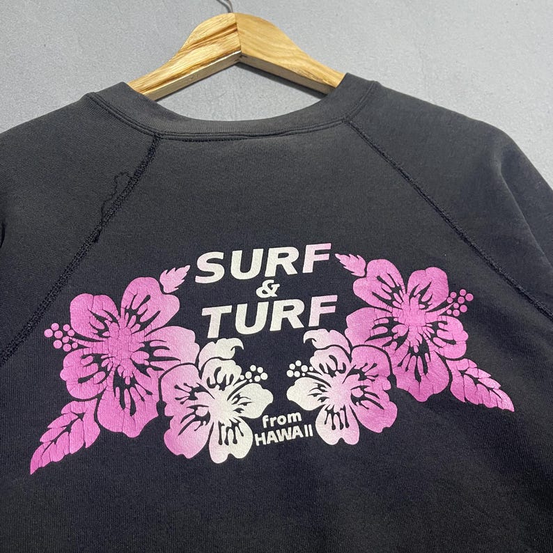 Op de afbeelding: Zwart sweatshirt met een bloemenontwerp in roze en wit. De tekst "SURF & TURF" is boven de bloemen gebogen, met "from HAWAII" eronder. Het sweatshirt hangt aan een houten hanger.
