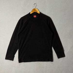 Puede incluir: Una camiseta negra de manga larga con cuello alto. La prenda está colgada de una percha de madera. Un pequeño logotipo rojo es visible en la parte superior del pecho. La camiseta está hecha de un material suave y cómodo.