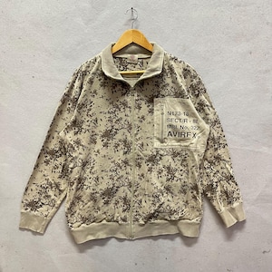 Puede incluir: Chaqueta con cremallera de camuflaje beige y marr&oacute;n con un peque&ntilde;o bolsillo en el pecho. El bolsillo tiene el texto "N173-18 SECT/R - 8L MRL No. 322 AVIREX". La chaqueta est&aacute; colgada de una percha de madera.