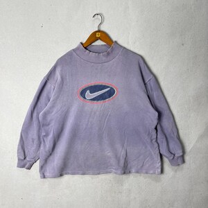 May include: A vintage lavender Nike sweatshirt with a mock neck. The sweatshirt features a large oval logo on the front with a blue swoosh, a pink outline, and a navy blue oval border. The long-sleeved top is hanging on a wooden hanger.