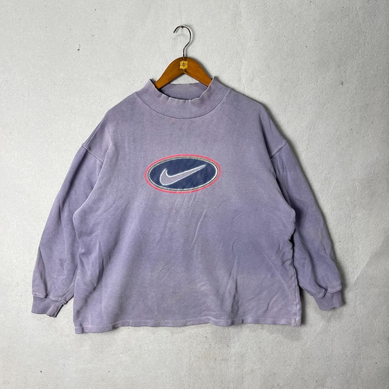May include: A vintage lavender Nike sweatshirt with a mock neck. The sweatshirt features a large oval logo on the front with a blue swoosh, a pink outline, and a navy blue oval border. The long-sleeved top is hanging on a wooden hanger.