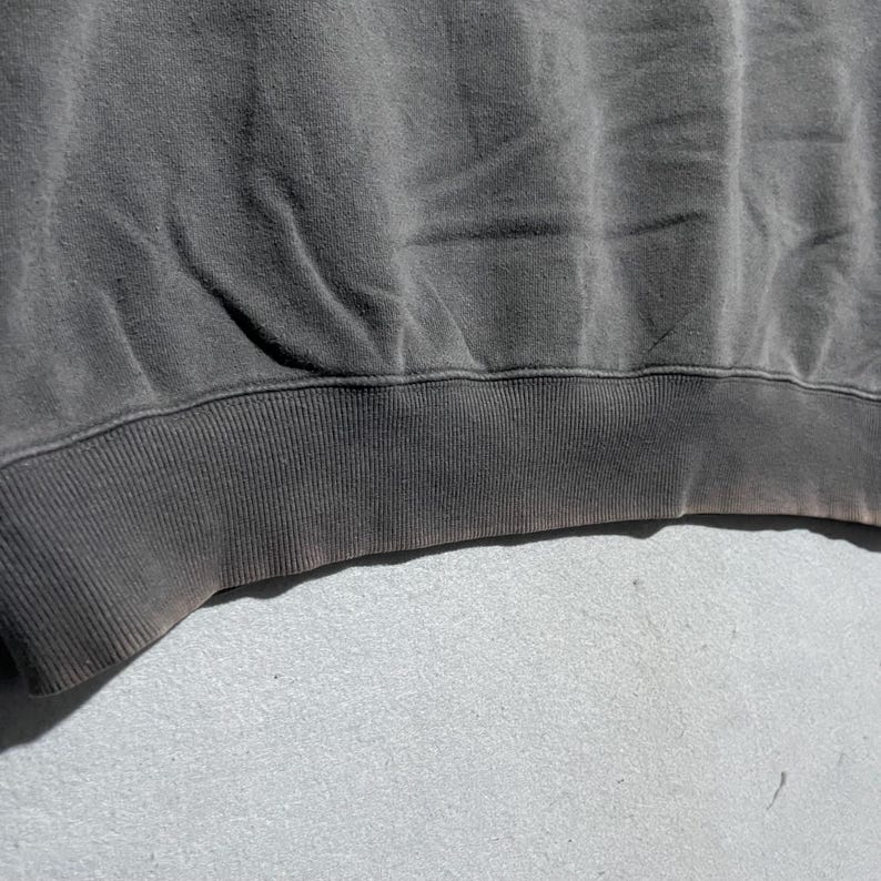 Puede incluir: Primer plano de una sudadera gris oscuro. El dobladillo inferior y los pu&ntilde;os son acanalados. La tela tiene un aspecto desgastado con sutiles variaciones de color, lo que sugiere que ha sido lavada y usada.