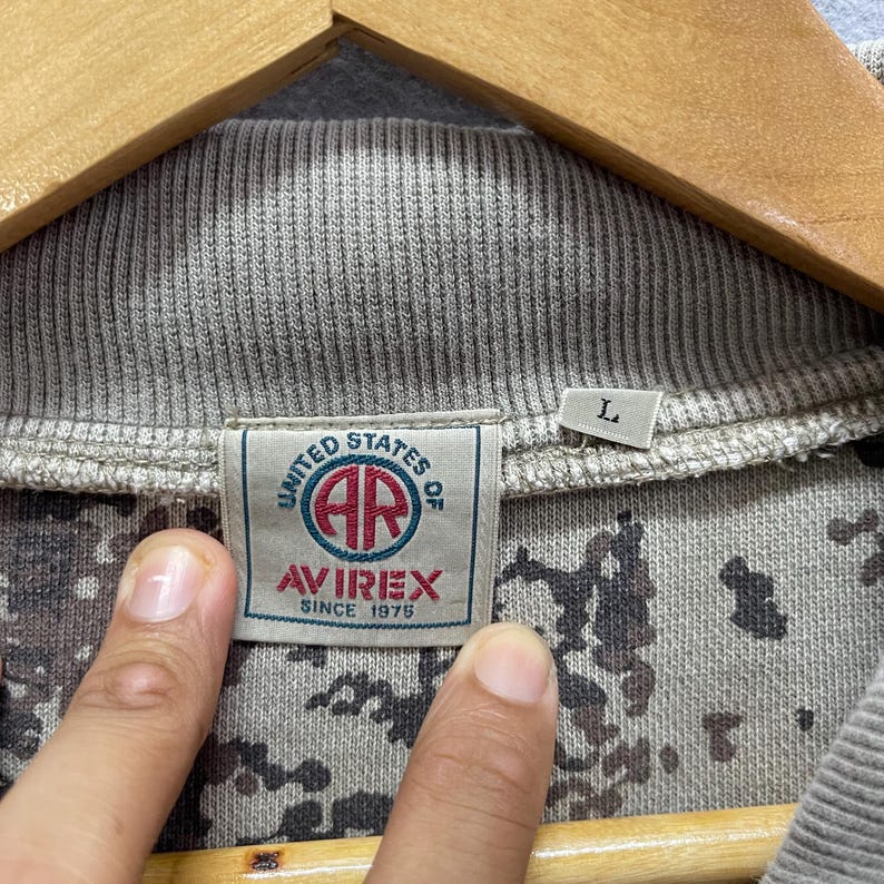 Puede incluir: Primer plano de una sudadera gris con estampado de camuflaje. La etiqueta dice "UNITED STATES OF AR AVIREX SINCE 1975". La sudadera tiene un cuello acanalado y una etiqueta de talla "L".