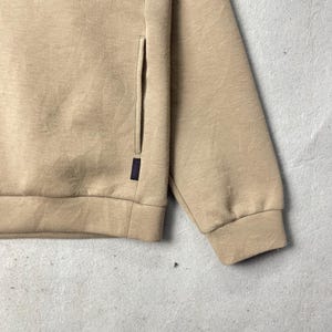 May include: Close-up of a tan sweatshirt with a side pocket and a dark purple tag. The sweatshirt has a ribbed cuff and hem. The fabric appears to be a soft, knit material. The background is a mottled gray.