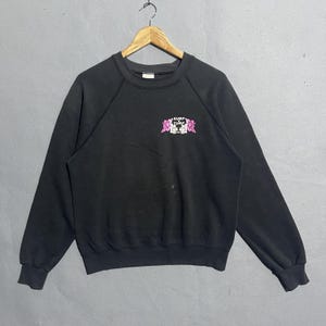 Op de afbeelding: Zwart sweatshirt met ronde hals met de tekst "Surf Tune" in wit en roze, samen met bloemenprints. Het sweatshirt hangt aan een houten hanger tegen een grijze achtergrond.