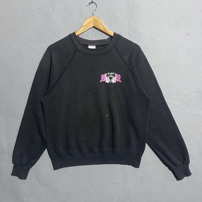 Op de afbeelding: Zwart sweatshirt met ronde hals met de tekst "Surf Tune" in wit en roze, samen met bloemenprints. Het sweatshirt hangt aan een houten hanger tegen een grijze achtergrond.