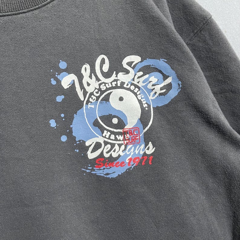 Puede incluir: Una sudadera gris con un dise&ntilde;o circular que muestra "T&C Surf Designs" y "Since 1971." El dise&ntilde;o incorpora un s&iacute;mbolo de yin-yang y "Hawaii Surf." El gr&aacute;fico es en blanco, azul y rojo.