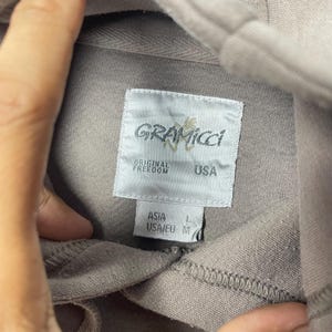 May include: Close-up of a taupe-colored garment tag with the brand name "Gramicci" in a stylized font. The tag also includes the text "Original Freedom" and "USA". The tag indicates sizes for Asia and USA/EU.
