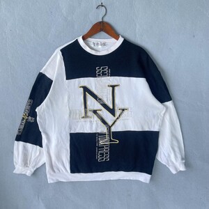 Könnte beinhalten: Ein Vintage-Sweatshirt mit einem Farbblock-Design in Weiß, Marineblau und Schwarz. Auf der Vorderseite befindet sich eine große goldene "NY"-Grafik mit zusätzlichem Text. Das Sweatshirt hängt an einem Holzbügel.