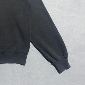 Op de afbeelding: Close-up van een zwarte sweatshirtmouw. De mouw is gemaakt van een zacht materiaal en heeft een geribde manchet. Het sweatshirt lijkt versleten.