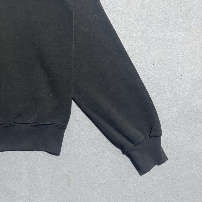 Op de afbeelding: Close-up van een zwarte sweatshirtmouw. De mouw is gemaakt van een zacht materiaal en heeft een geribde manchet. Het sweatshirt lijkt versleten.