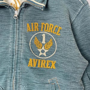 Peut inclure: Gros plan sur une veste en jean bleu sarcelle avec une fermeture &eacute;clair. La veste porte les mots "AIR FORCE" au-dessus d'un &eacute;cusson circulaire avec une &eacute;toile, des ailes et le chiffre "1". Le mot "AVIREX" est en dessous, en lettres dor&eacute;es.