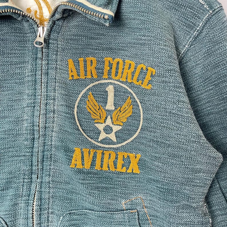 Peut inclure: Gros plan sur une veste en jean bleu sarcelle avec une fermeture &eacute;clair. La veste porte les mots "AIR FORCE" au-dessus d'un &eacute;cusson circulaire avec une &eacute;toile, des ailes et le chiffre "1". Le mot "AVIREX" est en dessous, en lettres dor&eacute;es.