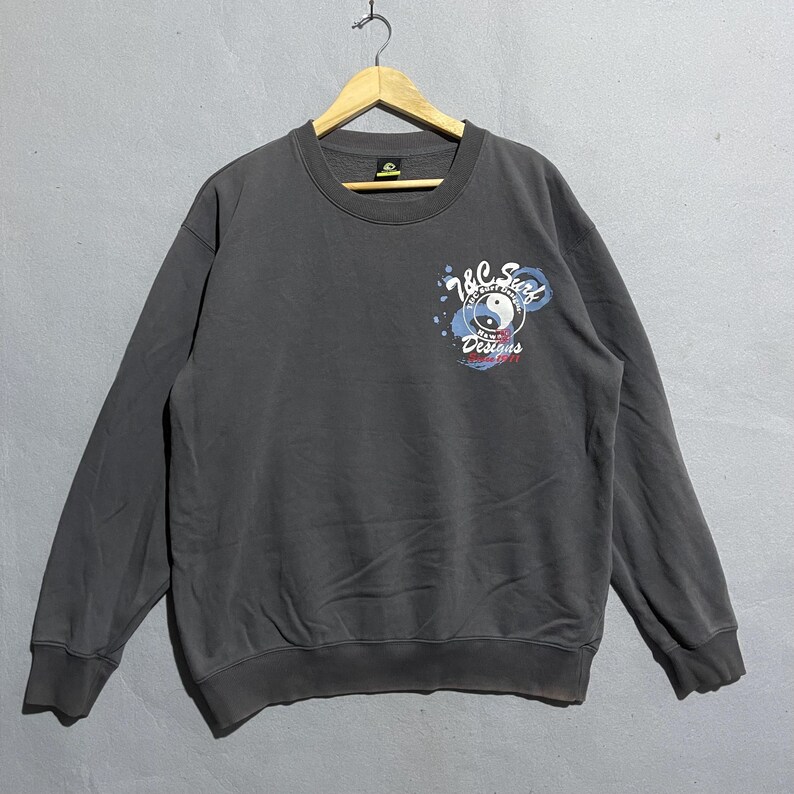 Puede incluir: Sudadera gris oscuro de cuello redondo con un gr&aacute;fico en la parte delantera. El gr&aacute;fico incluye el texto "Y&C Surf Designs" y un s&iacute;mbolo de yin-yang. La sudadera est&aacute; colgada en una percha de madera.