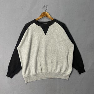 Könnte beinhalten: Ein graues und schwarzes Raglan-Sweatshirt hängt an einem Holzbügel. Das Sweatshirt hat einen grauen Körper mit schwarzen Ärmeln und ein schwarzes Dreieckdetail am Ausschnitt. Die Bündchen und der Ausschnitt sind gerippt.