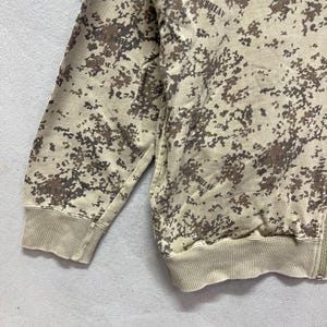 Puede incluir: Una sudadera con cremallera de camuflaje marr&oacute;n y beige. La sudadera presenta un patr&oacute;n de peque&ntilde;as formas irregulares en tonos marrones y beige. Los pu&ntilde;os y la cintura son de color beige liso. La cremallera es visible en el lado derecho.