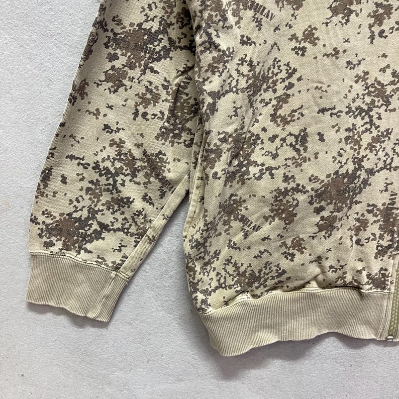 Puede incluir: Una sudadera con cremallera de camuflaje marr&oacute;n y beige. La sudadera presenta un patr&oacute;n de peque&ntilde;as formas irregulares en tonos marrones y beige. Los pu&ntilde;os y la cintura son de color beige liso. La cremallera es visible en el lado derecho.