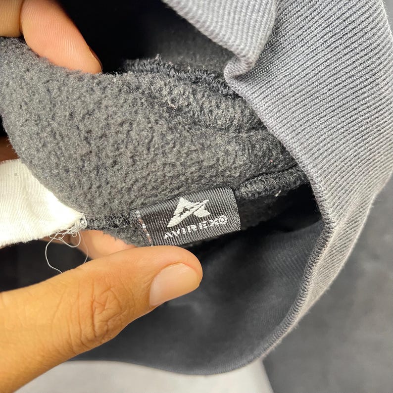 Puede incluir: Primer plano del interior de una sudadera gris oscuro con una etiqueta. La etiqueta es negra con texto blanco y un logotipo que dice "AVIREXO". La tela de la sudadera es un material suave y texturizado.