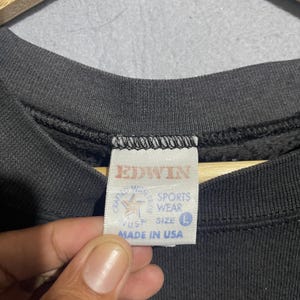 Op de afbeelding: Close-up van een zwart Edwin sweatshirt-label. Het witte label heeft rode tekst met "EDWIN" en "SPORTS WEAR". Het label vermeldt ook "MADE IN USA" en heeft maat "L".