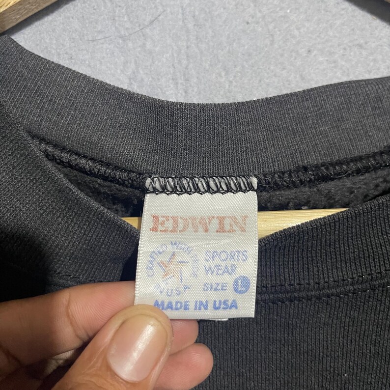 Op de afbeelding: Close-up van een zwart Edwin sweatshirt-label. Het witte label heeft rode tekst met "EDWIN" en "SPORTS WEAR". Het label vermeldt ook "MADE IN USA" en heeft maat "L".