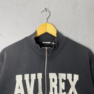 Puede incluir: Una sudadera negra con cremallera y el texto blanco "AVI REX" en el pecho. La sudadera tiene un cuello alto y una cremallera plateada. La prenda est&aacute; colgada de una percha de madera.