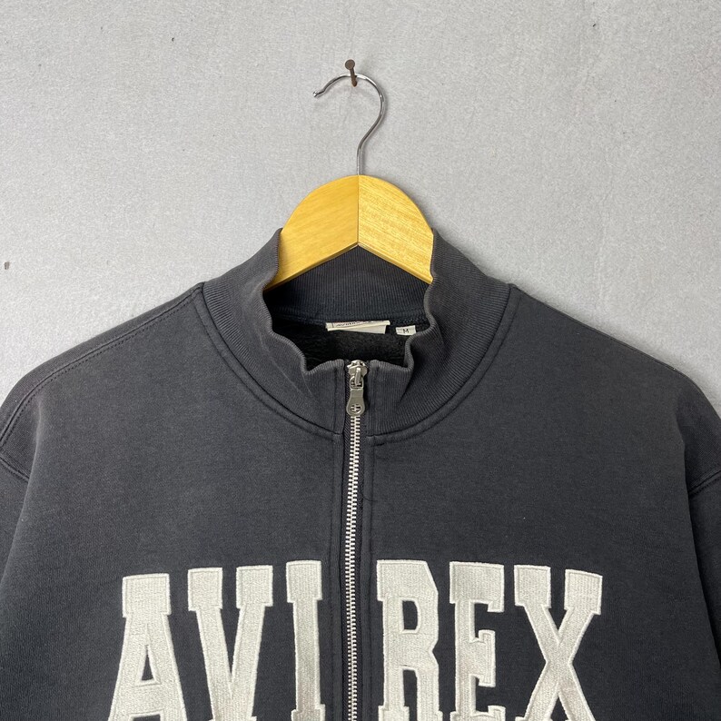 Puede incluir: Una sudadera negra con cremallera y el texto blanco "AVI REX" en el pecho. La sudadera tiene un cuello alto y una cremallera plateada. La prenda est&aacute; colgada de una percha de madera.