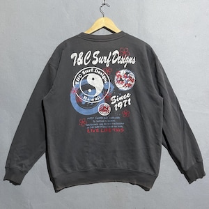 Puede incluir: Sudadera gris oscura con un dise&ntilde;o en la espalda que dice "T&C Surf Designs Hawaii Since 1971". El dise&ntilde;o incluye un s&iacute;mbolo de yin-yang, flores y patrones de olas. La sudadera est&aacute; colgada en una percha de madera.
