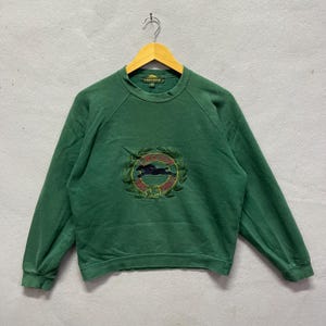 Puede incluir: Sudadera verde de cuello redondo con el logotipo de Longchamp bordado en la parte delantera. El logotipo presenta un caballo dentro de una corona de laurel. La sudadera está colgada en una percha de madera.