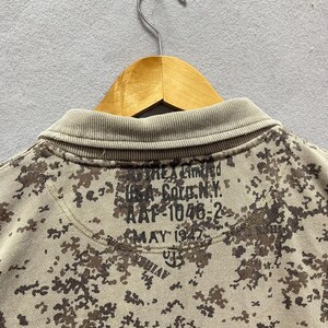Puede incluir: Primer plano de un polo de camuflaje en beige y marr&oacute;n. La camisa tiene un cuello acanalado y un patr&oacute;n de manchas marrones y beige. La parte posterior de la camisa tiene el texto "THE Limited", "164 Soto N.Y.", "AAF-1046-2" y "MAY 1947".