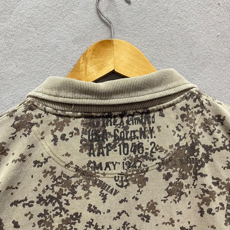 Puede incluir: Primer plano de un polo de camuflaje en beige y marr&oacute;n. La camisa tiene un cuello acanalado y un patr&oacute;n de manchas marrones y beige. La parte posterior de la camisa tiene el texto "THE Limited", "164 Soto N.Y.", "AAF-1046-2" y "MAY 1947".