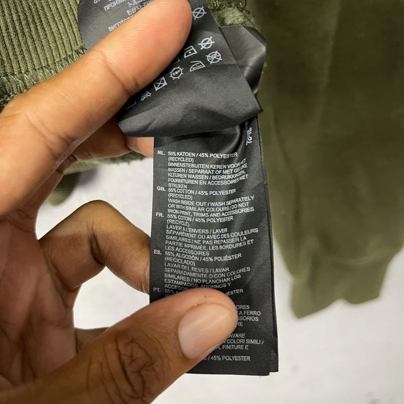 Puede incluir: Primer plano de una etiqueta de ropa negra con texto blanco y s&iacute;mbolos de cuidado. La etiqueta, sujeta por una mano, est&aacute; adherida a una prenda verde. Proporciona instrucciones de lavado y la composici&oacute;n del tejido.