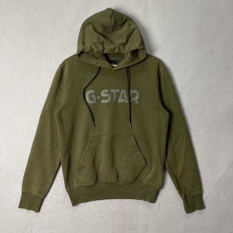 Puede incluir: Sudadera con capucha verde oliva con el logotipo de G-Star en el pecho. La sudadera tiene una capucha con cord&oacute;n y un bolsillo de canguro. La tela parece ser un material suave y de peso medio.