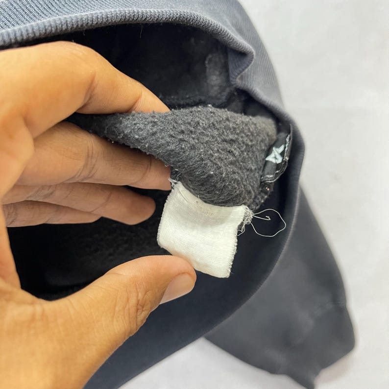 Puede incluir: Primer plano del interior de una sudadera gris oscuro con una etiqueta blanca. El interior es de un material gris m&aacute;s oscuro y difuso. La etiqueta es blanca y rectangular. Una mano sostiene la sudadera.