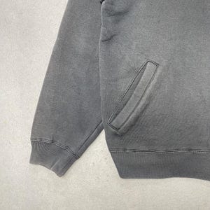 Puede incluir: Una sudadera gris carb&oacute;n con bolsillo de canguro y pu&ntilde;os y dobladillo acanalados. La prenda tiene un aspecto descolorido, lo que sugiere una est&eacute;tica vintage o desgastada. La tela parece ser un material suave y c&oacute;modo.