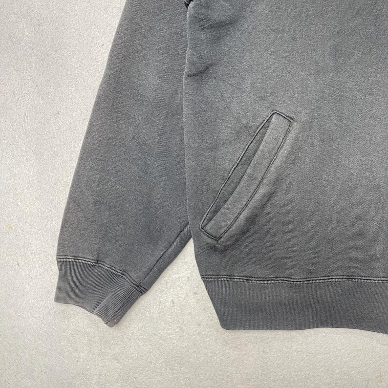 Puede incluir: Una sudadera gris carb&oacute;n con bolsillo de canguro y pu&ntilde;os y dobladillo acanalados. La prenda tiene un aspecto descolorido, lo que sugiere una est&eacute;tica vintage o desgastada. La tela parece ser un material suave y c&oacute;modo.