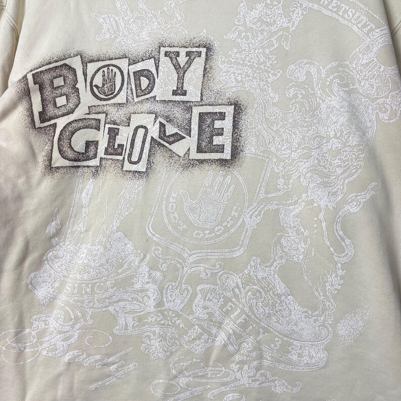 Puede incluir: Una sudadera de color crema con un gr&aacute;fico grande de estilo pintura en aerosol marr&oacute;n oscuro que dice BODY GLOVE. Debajo del texto hay un estampado blanco intrincado de estilo her&aacute;ldico con un logotipo de mano, volutas y el texto SINCE FIFTY THREE.